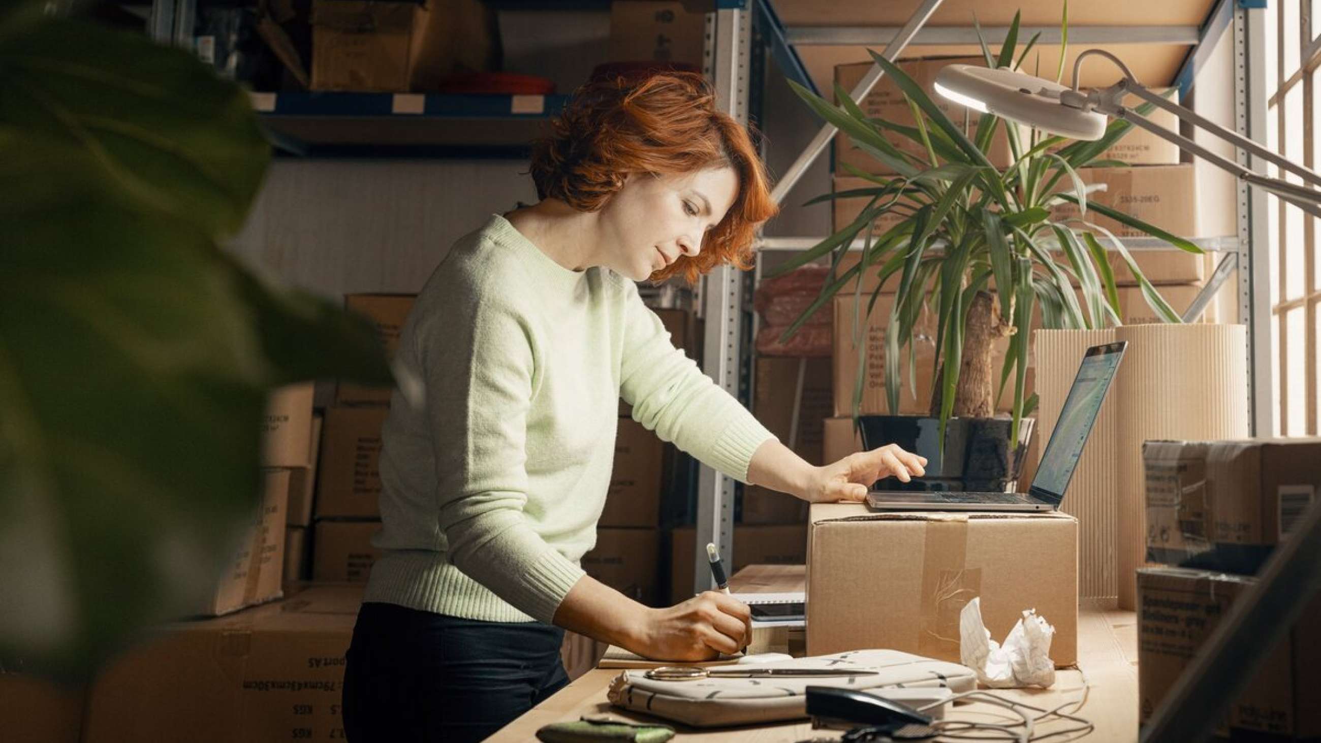 A female online store owner stands by a cardboard box on a work table with many drawers in the background. She takes notes in a pad while holding the other hand on an open laptop.