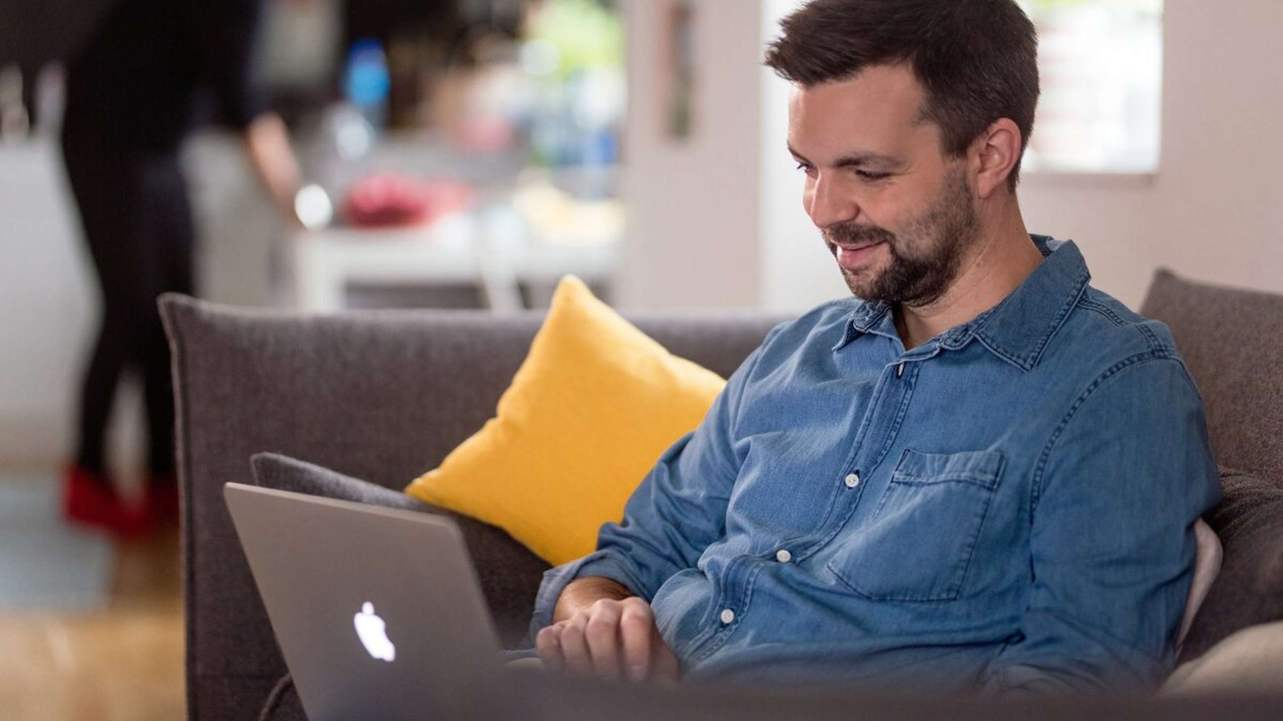 Young man with beard and denim shirt sitting on the sofa and smiling with a laptop in his lap.